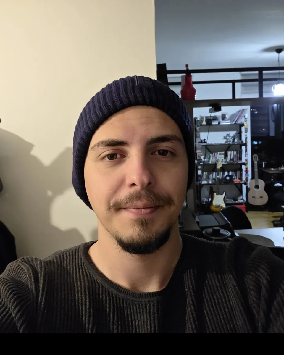 Portrait of Kürşat Öztürk in a dark knit beanie and sweater, at home with guitars on a shelf behind him.